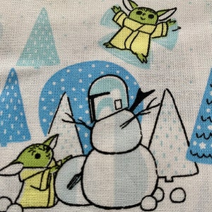 May include: A white fabric with a blue and white winter scene featuring a snowman with a Mandalorian helmet, two green alien characters, and blue and white trees.