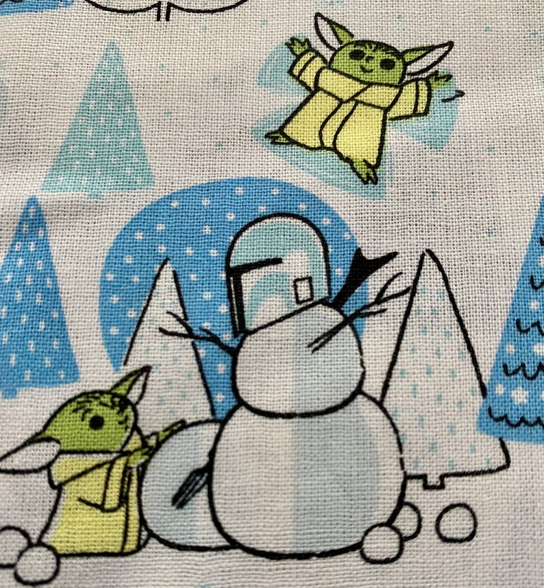 May include: A white fabric with a blue and white winter scene featuring a snowman with a Mandalorian helmet, two green alien characters, and blue and white trees.