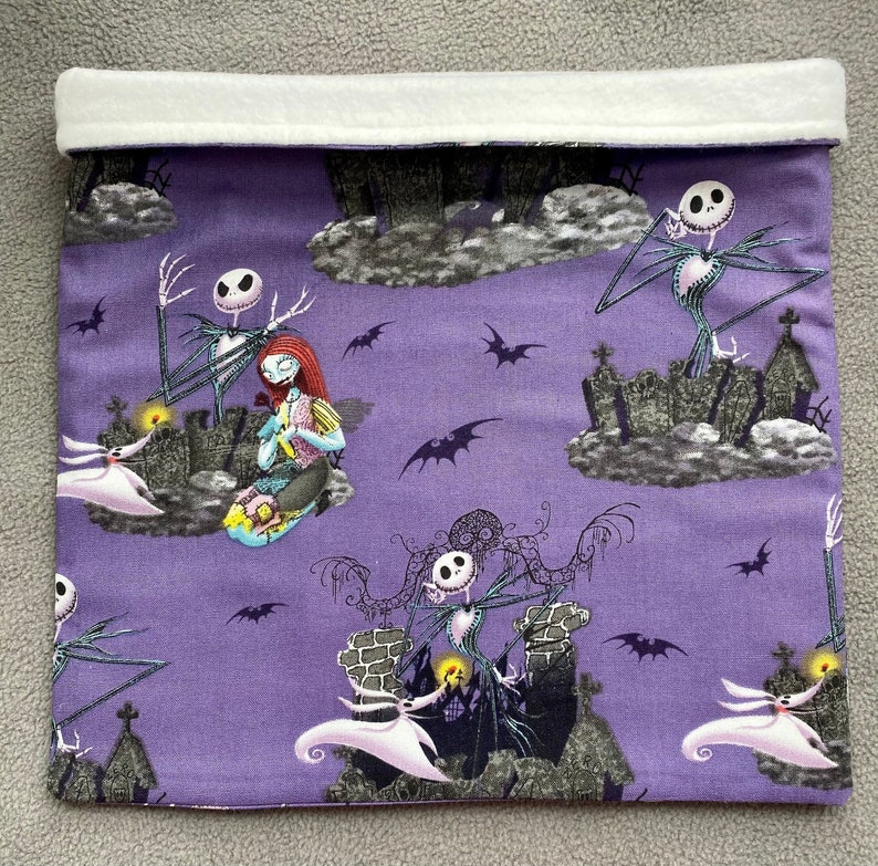 May include: Purple fabric pouch with a white fleece lining. The pouch features a repeating pattern of Jack Skellington and Sally from the Nightmare Before Christmas movie. The characters are depicted in various scenes from the movie, including Jack Skellington standing on a hilltop and Sally sitting on a cloud.