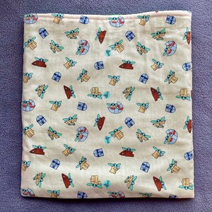 May include: A beige fabric square pillowcase with a repeating pattern of small green and blue cartoon characters. The characters are all facing forward and have large eyes and ears. The background is a light beige color.