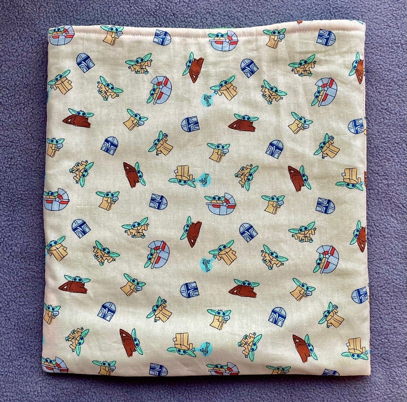 May include: A beige fabric square pillowcase with a repeating pattern of small green and blue cartoon characters. The characters are all facing forward and have large eyes and ears. The background is a light beige color.
