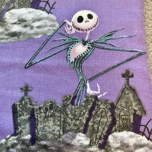 May include: A purple fabric with a white and black illustration of Jack Skellington from The Nightmare Before Christmas. He is standing on a graveyard with a church and tombstones.
