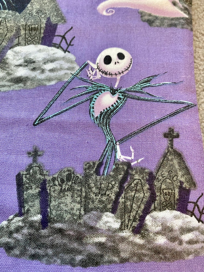 May include: A purple fabric with a white and black illustration of Jack Skellington from The Nightmare Before Christmas. He is standing on a graveyard with a church and tombstones.