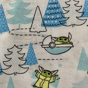 May include: A white fabric with a repeating pattern of blue and green Christmas trees and a green character from the Star Wars franchise. The character is wearing a brown robe and is flying in a spaceship. The character is also standing on the ground.