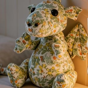 May include: A floral patterned dragon soft toy in green, yellow, and orange. The dragon has large black eyes, small ears, and wings. The soft toy is sitting upright.