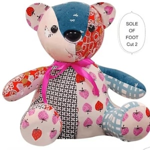 May include: A patchwork teddy bear constructed from various fabric patterns including pink strawberries, black and white geometric shapes, and blue textures. The bear features a bright pink ribbon tied around its neck and black button eyes.