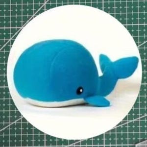May include: A soft, plush blue whale toy with a white underside, a black eye, and a curved tail fin. The whale is lying down on a white surface, against a green grid. The toy is designed to be soft and cuddly.