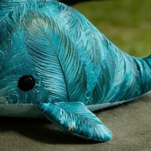 May include: A teal and blue plush whale toy with a feather-like design. The toy has a rounded body, a small tail, and fins. It features a black eye and is resting on a green surface.