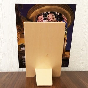 Może przedstawiać: Drewniana ramka na zdjęcia ze stojakiem, wykonana z jasnego drewna. Ramka zawiera zdjęcie trzech uśmiechniętych os&oacute;b.