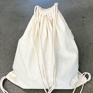 Puede incluir: Una bolsa de cordón blanca lisa hecha de tela de algodón. La bolsa está vacía y tiene una parte superior fruncida con un cierre de cordón.