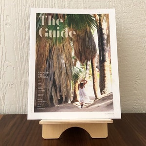Może przedstawiać: Okładka czasopisma z tytułem "The Guide" i tekstem "Greater Palm Springs". Okładka przedstawia zdjęcie osoby idącej przez gaj palmowy.