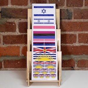 A wooden display stand with three 5x7 sticker cards featuring different flags. The top card shows the flag of Israel, the middle card shows the transgender pride flag, and the bottom card shows the flag of British Columbia.