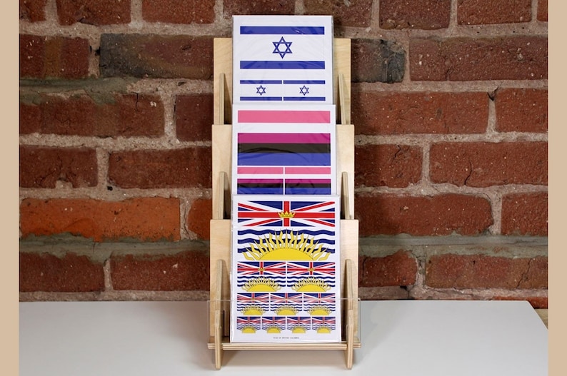 A wooden display stand with three 5x7 sticker cards featuring different flags. The top card shows the flag of Israel, the middle card shows the transgender pride flag, and the bottom card shows the flag of British Columbia.
