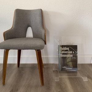 May include: A gray upholstered chair with wooden legs and a clear acrylic magazine rack holding a book titled "Building Conversion & Renovation".