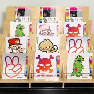 A wooden display stand with twelve sections each featuring various stickers designs, including a skull, a peace sign, a monster, and a cat. The stand has a clear acrylic front allow full view of the stickers.