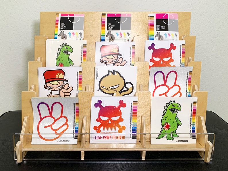 A wooden display stand with twelve sections each featuring various stickers designs, including a skull, a peace sign, a monster, and a cat. The stand has a clear acrylic front allow full view of the stickers.