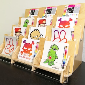 A wooden 12 pocket display stand with four shelves for showcasing stickers. The stickers feature various colorful designs, including a skull, a monster, and a peace sign. The stand is made of light birch wood and has a clear acrylic front.