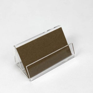 May include: A clear acrylic business card holder with a brown card insert. The holder is angled to display the card.