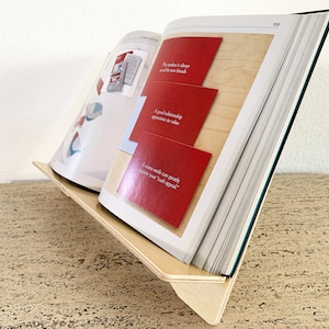 May include: A wooden book stand holding an open book with red and white pages. The book is propped up at an angle, displaying text on the pages. The stand is made of light-colored wood.