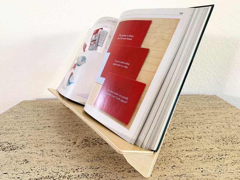 Side-angle view of a natural birch plywood 18-inch book display stand holding an open, multi-page catalog on a wooden table against a white background.