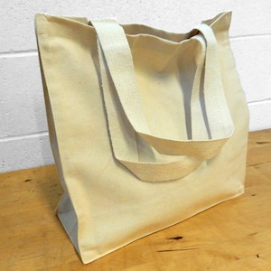 May include: A beige canvas tote bag with sturdy handles. The bag is rectangular with a flat bottom and is sitting on a wooden surface. The background is a white brick wall. The bag is empty.