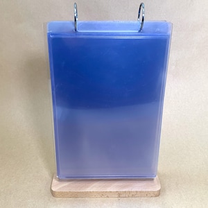 May include: A clear, blue plastic display stand with a wooden base. The stand has a rectangular shape and two silver metal rings at the top. The blue color fades from dark at the top to light at the bottom.