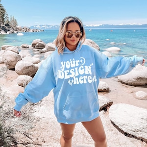 K&ouml;nnte beinhalten: Ein hellblauer Hoodie mit dem Text "your design here" in Wei&szlig;. Der Hoodie hat ein Mond- und Sternen-Motiv. Die Person, die den Hoodie tr&auml;gt, steht an einem Strand mit Felsen und einem See im Hintergrund.