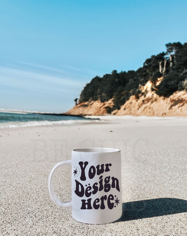 May include: White ceramic mug with the text "Your Design Here" in a retro font, accented with stars. The mug is on a sandy beach with the ocean and a cliff in the background. The sky is blue.