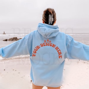 May include: A light blue hoodie with the text "YOUR DESIGN HERE" in orange on the back. The hoodie is worn by a person standing on a white sandy beach.