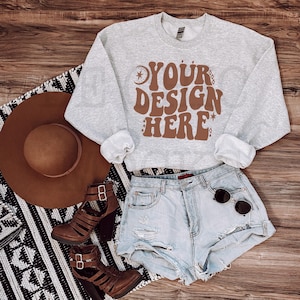 Puede incluir: Una sudadera gris recortada con el texto "YOUR DESIGN HERE" en letras marrones. La sudadera se combina con pantalones cortos de mezclilla, botas marrones y un sombrero marrón.