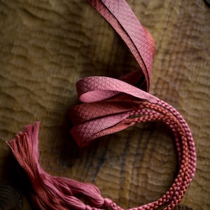 Puede incluir: Primer plano de un cord&oacute;n trenzado rojo y rosa con borlas. El cord&oacute;n est&aacute; sobre una superficie de madera.