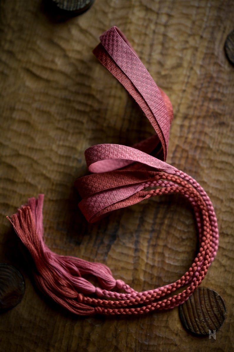 Puede incluir: Primer plano de un cord&oacute;n trenzado rojo y rosa con borlas. El cord&oacute;n est&aacute; sobre una superficie de madera.