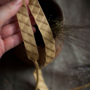 Puede incluir: Primer plano de una cinta de color dorado con un patr&oacute;n de diamante. La cinta se sostiene en una mano y est&aacute; anudada.