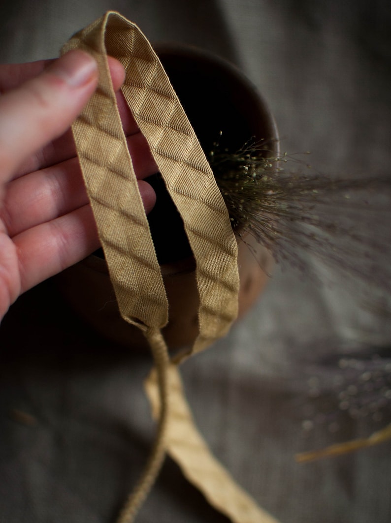 Puede incluir: Primer plano de una cinta de color dorado con un patr&oacute;n de diamante. La cinta se sostiene en una mano y est&aacute; anudada.