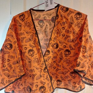 May include: Orange and black fabric wrap with a skull and bird print. The wrap has a V-neck and is trimmed with black fabric. The tag says "Size 18-20".