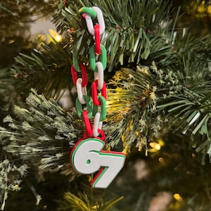 May include: A Christmas tree decoration with a chain of red, white, and green links. The ornament has the number 67 in white with a red outline. It is hanging on a green Christmas tree.