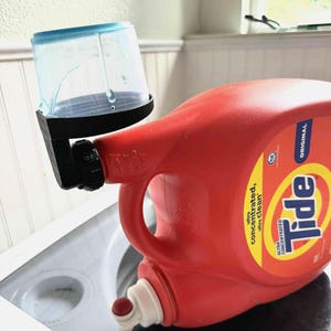 May include: A red Tide laundry detergent bottle with a black and blue measuring cup attached. The bottle is labeled "Original" and "Ultra Concentrated, Ultra Clean". The measuring cup is clear with blue accents.
