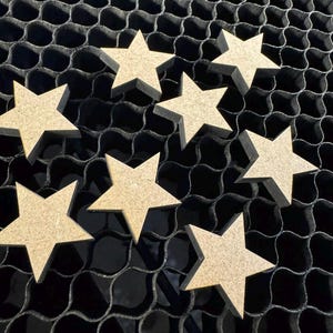 May include: A collection of unfinished wooden star cutouts. The stars are light brown and have a five-point design. They are arranged on a black honeycomb-patterned surface, creating a contrast between the natural wood and the dark background.