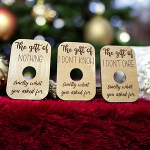 May include: Three wooden gift tags with humorous phrases: "The gift of NOTHING," "The gift of I DON'T KNOW," and "The gift of I DON'T CARE." Each tag has a circular hole and the text "Exactly what you asked for."