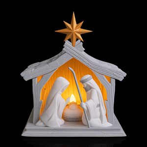May include: A white nativity scene with a gold star. The scene features Mary, Joseph, and baby Jesus within a wooden-style structure. The interior is illuminated with a warm glow, creating a peaceful holiday display.