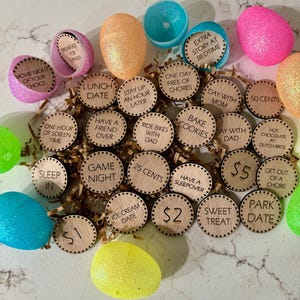 May include: Wooden tokens with various activities and rewards written on them, such as "Movie Night of Your Choice", "Lunch Date", "Game Night", "Ice Cream Date", "Have a Sleepover", "Bake Cookies", "Ride Bikes with Dad", "One Hour of Screen Time", "Stay Up an Hour Later", "Sleep In", "One Day Free of Chores", "Day with Mum", "Day with Dad", "50 Pence", "25 Pence", "£1", "£2", "£5", "Sweet Treat", "Park Date", "Get Out of a Chore", "Hot Chocolate at Dutch Bros", "Extra Story at Bedtime", and "Breakfast for Dinner". The tokens are scattered on a brown surface with several plastic Easter eggs.
