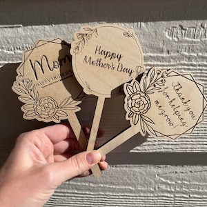 Könnte beinhalten: Drei Holz-Pflanzenmarker mit floralen Designs und gravierten Botschaften. Der erste Marker sagt "Mama Frohes Muttertags", der zweite sagt "Frohes Muttertags", und der dritte sagt "Danke, dass du mir beim Wachsen hilfst!"