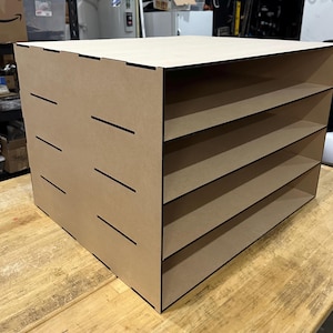 May include: A light brown wooden storage organizer featuring four horizontal open shelves. The side panel displays a series of horizontal slots, indicating a modular construction. The unit has a flat top surface and a rectangular frame design.