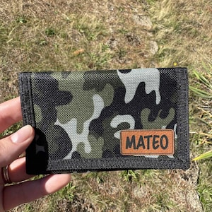 May include: A camouflage pattern wallet with a black border and a brown leather patch that reads "MATEO". The wallet is held against a backdrop of dry grass.