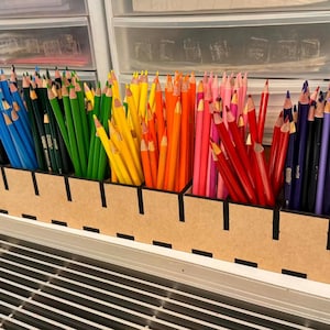 Op de afbeelding: Een houten potloodorganizer gevuld met een regenboog aan kleurpotloden. De potloden zijn gesorteerd op kleur, waaronder zwart, blauw, groen, geel, oranje, roze, rood, paars en bruin. De organizer staat op een wit oppervlak.