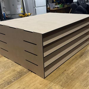May include: A light brown, rectangular desktop organizer made of flat, smooth panels. The unit features four horizontal open shelves for storage, with visible interlocking slot joints along the side panels and a clean, matte finish throughout.