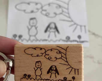Könnte beinhalten: Ein Holzschlüsselanhänger mit einem handgezeichneten Bild einer Familie mit Sonne und Wolken. Die Zeichnung ist in schwarzer Tinte auf einer hellbraunen Holzoberfläche.