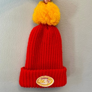 May include: A red knit beanie with a large yellow pom-pom on top. The beanie features a folded brim and a patch with the letters "USC" and a football graphic.