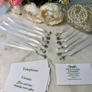 May include: An assortment of silver charms attached to white ribbons, including a crown, airplane, and Eiffel Tower. The charms are displayed with cards that read "Telephone" and "Crown, You will live happily ever after."