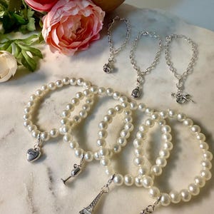 May include: Several pearl bracelets with silver charms, including a heart with "Love" engraved, a champagne glass, and a miniature Eiffel Tower. Also, three silver charm bracelets with various charms. Pink and white flowers are in the background.